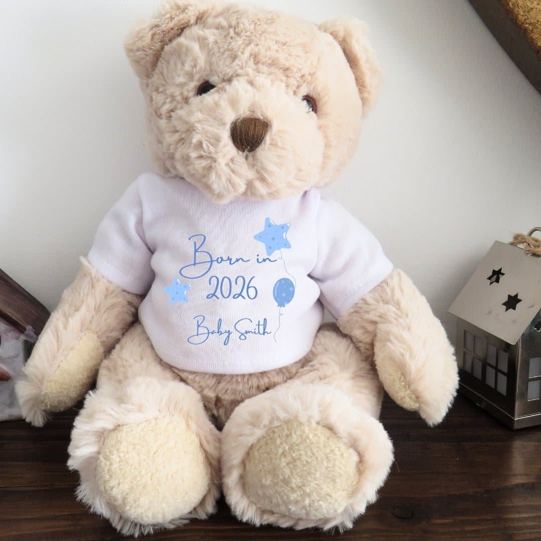 Teddy bear in a T-shirt printed Born in 2026 with blue lettering and blue balloons